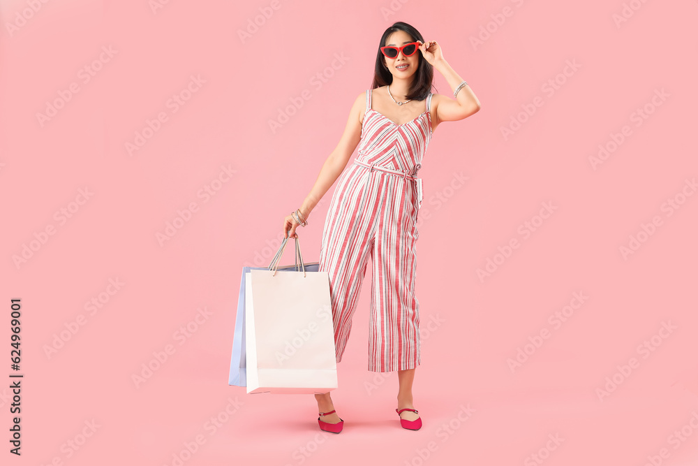 Happy beautiful young Asian woman in sunglasses with shopping bags on pink background Happy beautiful young Asian woman in sunglasses with shopping bags on pink background