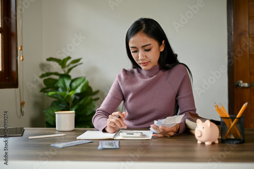 A serious Asian woman sitting at a table using a calculator, checking her electric bills