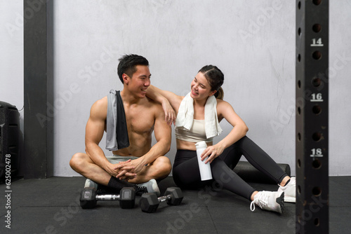 Young asian couple amidst workout on gym floor, surrounded by dumbbells, engaging in a moment of rest.