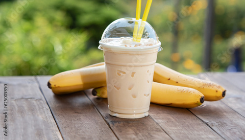Vászonkép banana milkshake in disposable plastic glass with straw on wooden table outdoors