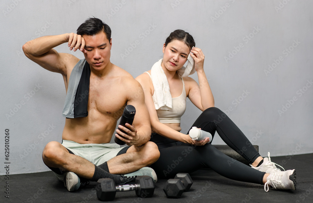 Asian strong young male muscular shirtless fitness model in sporty shorts and female athlete in sport bra sitting smiling taking break holding water bottle talking together in gym on gray background Asian strong young male muscular shirtless fitness model in sporty shorts and female athlete in sport bra sitting smiling taking break holding water bottle talking together in gym on gray background