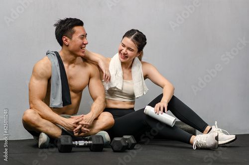 Asian strong young male muscular shirtless fitness model in sporty shorts and female athlete in sport bra sitting smiling taking break holding water bottle talking together in gym on gray background