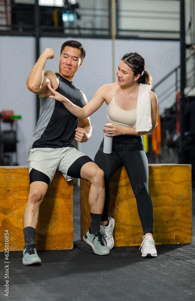 Asian strong young fit male muscular fitness athlete model showing off bicep muscle to female friend sitting take break drink water wipe sweat on wood box smiling hold thumb up complimenting in gym