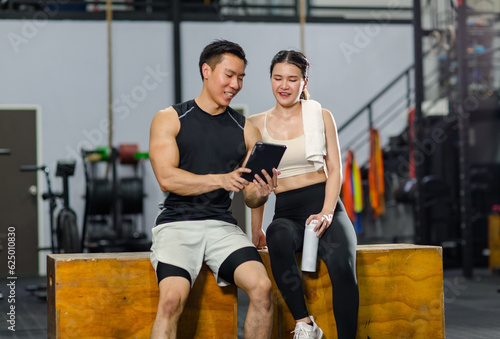 Asian strong young fit male muscular fitness athlete model and female friend sitting take break drink water wipe sweat on wood box smiling talking together showing schedule on tablet computer in gym