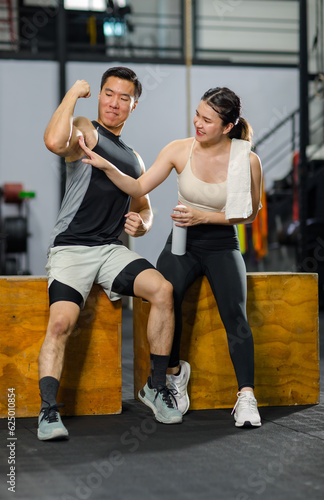Asian strong young fit male muscular fitness athlete model showing off bicep muscle to female friend sitting take break drink water wipe sweat on wood box smiling hold thumb up complimenting in gym