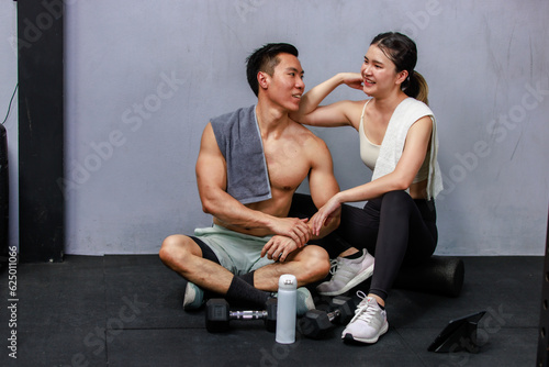 Asian strong young male muscular shirtless fitness model in sporty shorts and female athlete in sport bra sitting smiling taking break holding water bottle talking together in gym on gray background