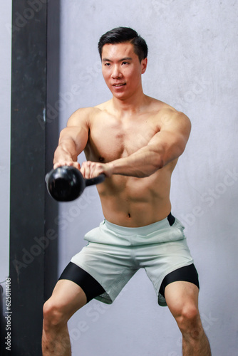 Asian strong handsome young male muscular shirtless fitness model in sporty shorts standing smiling holding lifting metal kettlebell dumbbell showing biceps triceps muscle in gym on gray background © Bangkok Click Studio