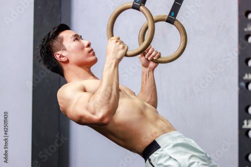 Asian strong handsome young shirtless male muscular fitness model in sporty shorts holding pushing up gymnastic rings lifting working out core muscle strength bodyweight in gym on gray background
