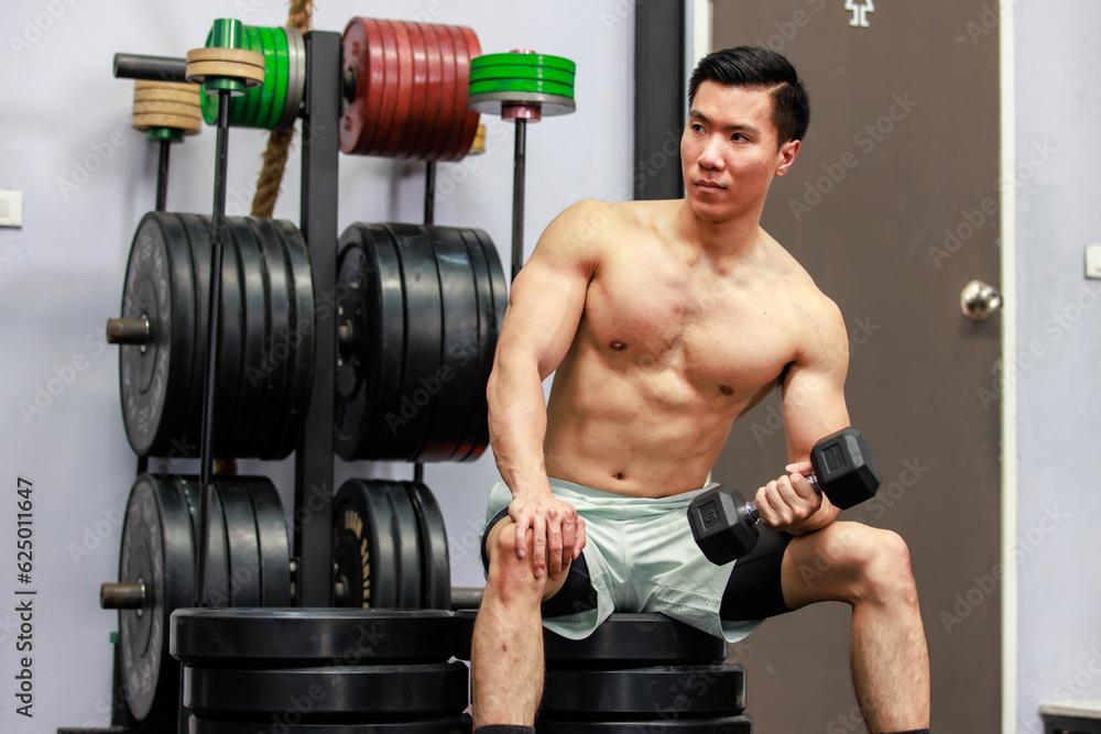 Millennial Asian fit strong handsome shirtless muscular male fitness model athlete in shorts sitting on barbell weights lifting training exercising alone in gym. Millennial Asian fit strong handsome shirtless muscular male fitness model athlete in shorts sitting on barbell weights lifting training exercising alone in gym.