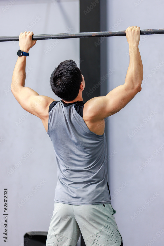 Naklejka premium Asian strong unrecognizable unknown young shirtless male muscular fitness model in sporty shorts holding pulling up gymnastic rings lifting working out back muscle strength in gym on gray background
