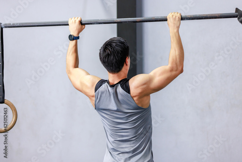 Asian strong unrecognizable unknown young shirtless male muscular fitness model in sporty shorts holding pulling up gymnastic rings lifting working out back muscle strength in gym on gray background