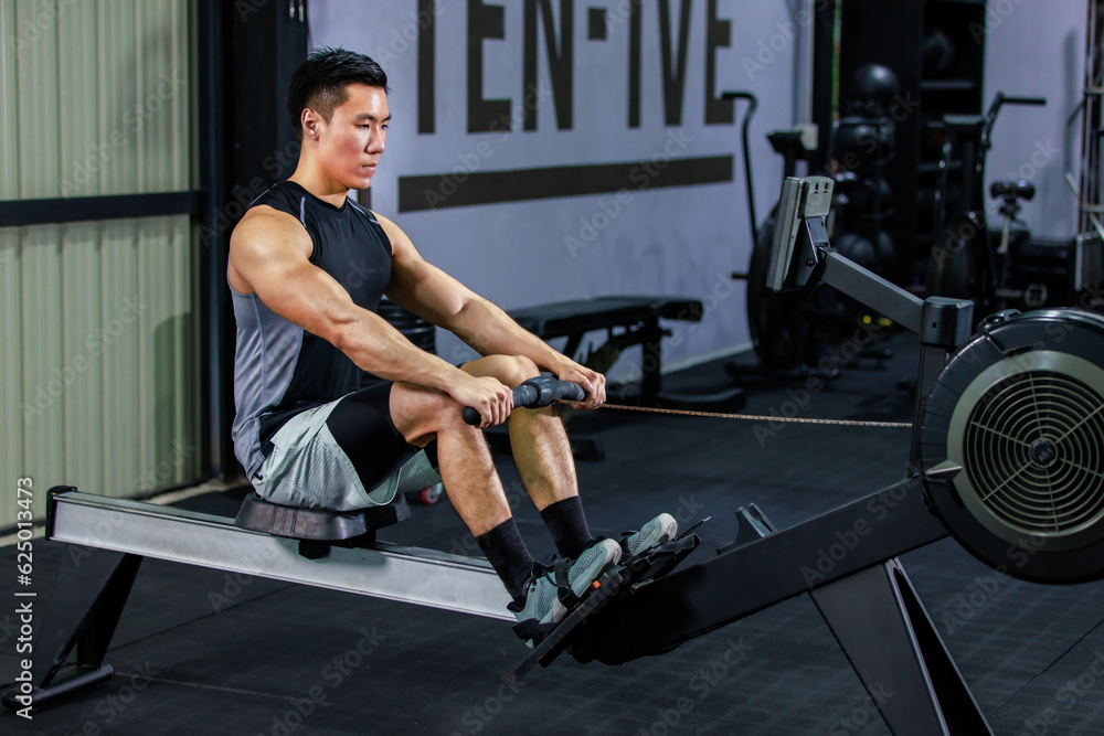 Asian strong young fit male muscular fitness athlete model in sleeveless shirt and sporty shorts sitting using heavy cable pulling machine working out training exercising in gym.