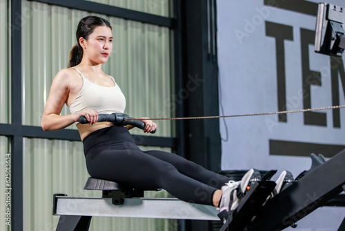 Asian strong young fit female muscular fitness athlete model in sexy sport bra and legging sitting looking at screen monitor using heavy cable pulling machine working out training exercising in gym