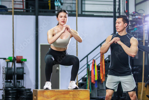 Asian strong young fit male trainer helping teaching female muscular fitness athlete model in sexy sport bra and legging training exercising squatting on wooden box in gym full of exercise equipment