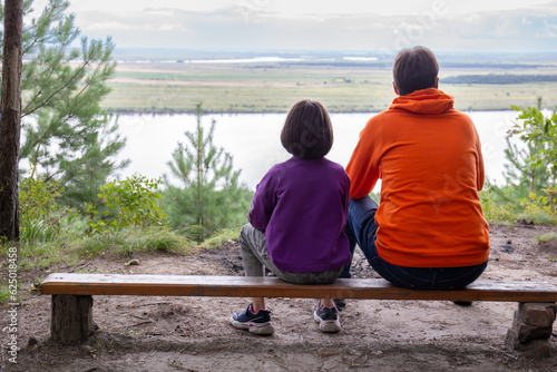 Dad and daughter sit on a bench with their backs to the camera and admire the beautiful natural landscape from above.