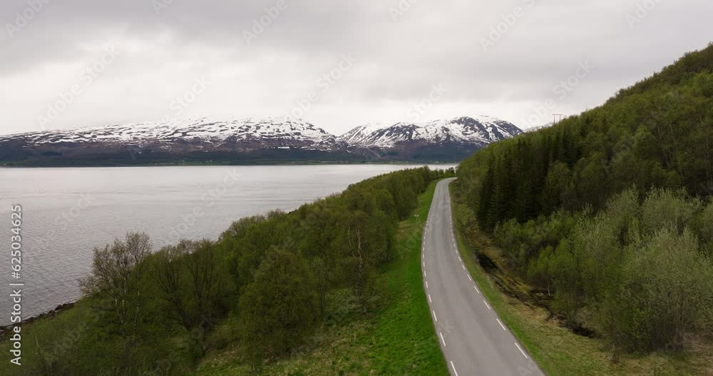 Landscape Of Unwinding Road Overlooking The Sea And Alps In Distance On A Cloudy Day In Northern Norway. aerial, wide shot Landscape Of Unwinding Road Overlooking The Sea And Alps In Distance On A Cloudy Day In Northern Norway. aerial, wide shot