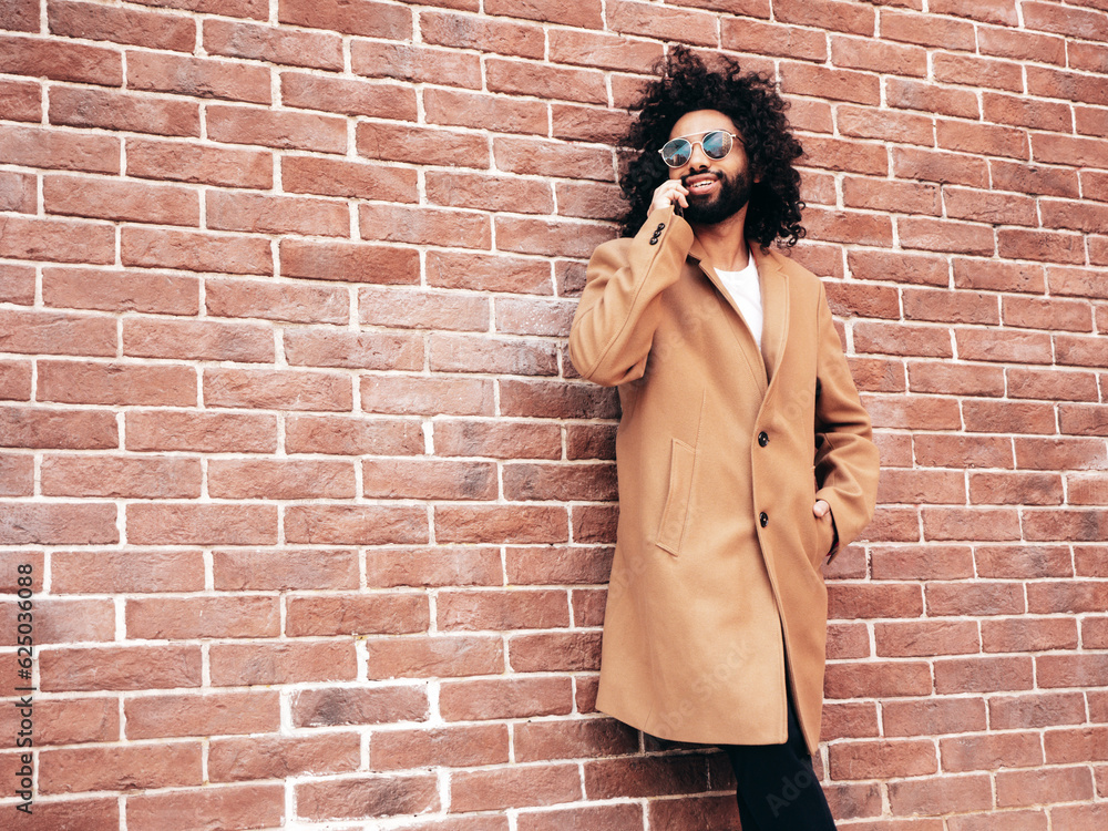 Handsome hipster model.  Unshaven Arabian man dressed in brown coat clothes. Fashion male with long curly hairstyle posing in street. Cheerful and happy. using smartphone, talks at mobile phone