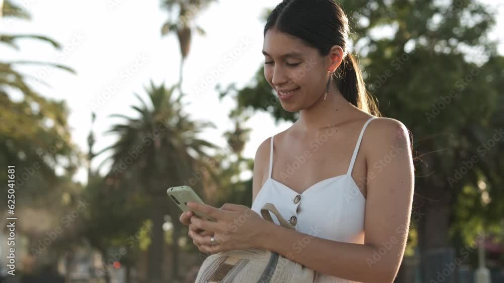 Low angle view of cheerful Latin American female smiling and checking messages on cellphone while standing in sunlit park Low angle view of cheerful Latin American female smiling and checking messages on cellphone while standing in sunlit park