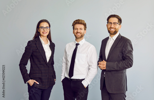 Obraz na plátně Portrait of a group of three young cheerful business people in suits standing confidently, looking at the camera and smiling isolated on a studio grey background