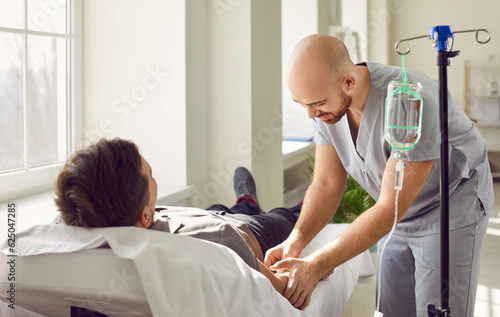 Nursing staff at clinic or hospital gives intravenous vitamin drip or medicine infusion to patient. Nurse personnel inserts venous IV line needle in arm vein of adult man lying on bed in medical ward photo