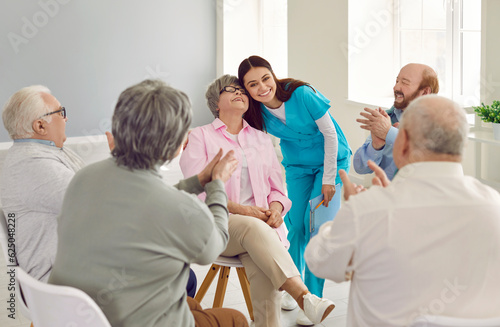 Happy young nurse and senior patients in the retirement home having fun together. Smiling girl caretaker hugs an old woman while other elderly people are clapping hands. Love, care, support concept