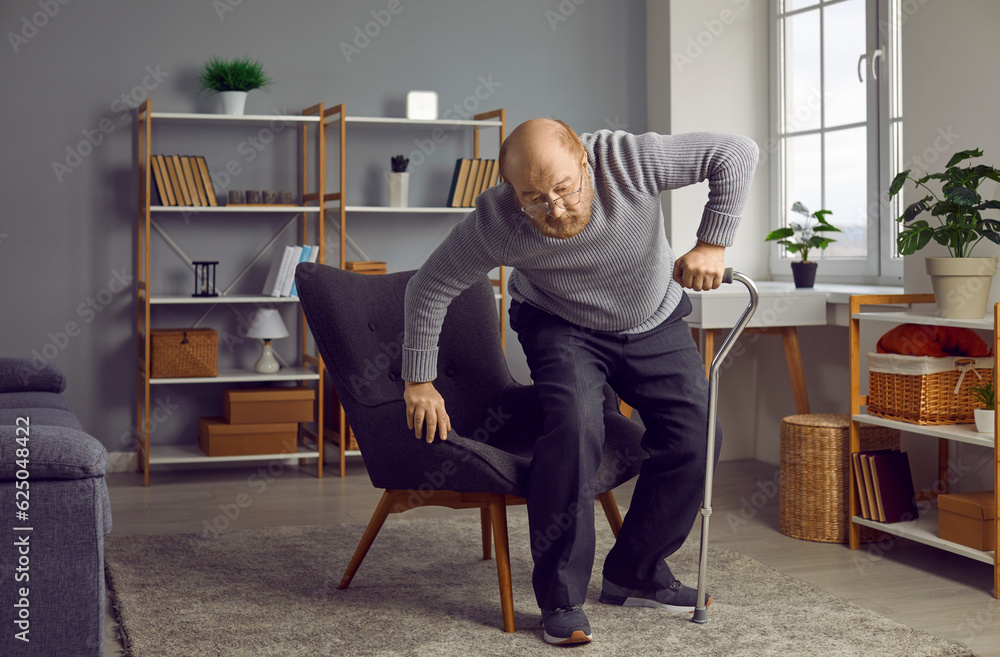 Retired old man with aching joints uses a cane to walk. Bald senior man with a walking stick stands up from an armchair in the living room at home