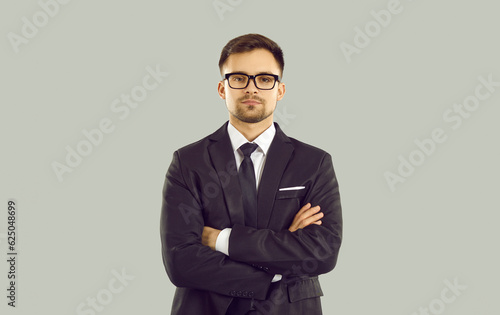 Studio portrait of serious young businessman. Handsome young European business man in classic suit, shirt, tie and glasses standing with his arms folded isolated on grey background