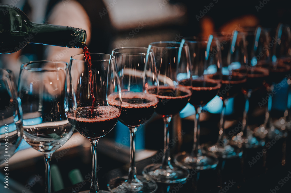 Party and celebration concepts. Bartender pours red wine in glasses at bar. Male sommelier pouring red wine into long-stemmed wineglasses. - obrazy, fototapety, plakaty 