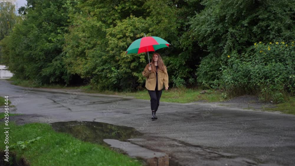 Wide shot confident charming woman with green and red umbrella walking in slow motion on rainy day outdoors. Portrait of beautiful brunette Caucasian lady strolling on suburban road outdoors Wide shot confident charming woman with green and red umbrella walking in slow motion on rainy day outdoors. Portrait of beautiful brunette Caucasian lady strolling on suburban road outdoors