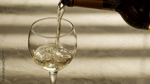 White wine from a bottle is poured into a wine glass illuminated by the sun through the blinds on the window