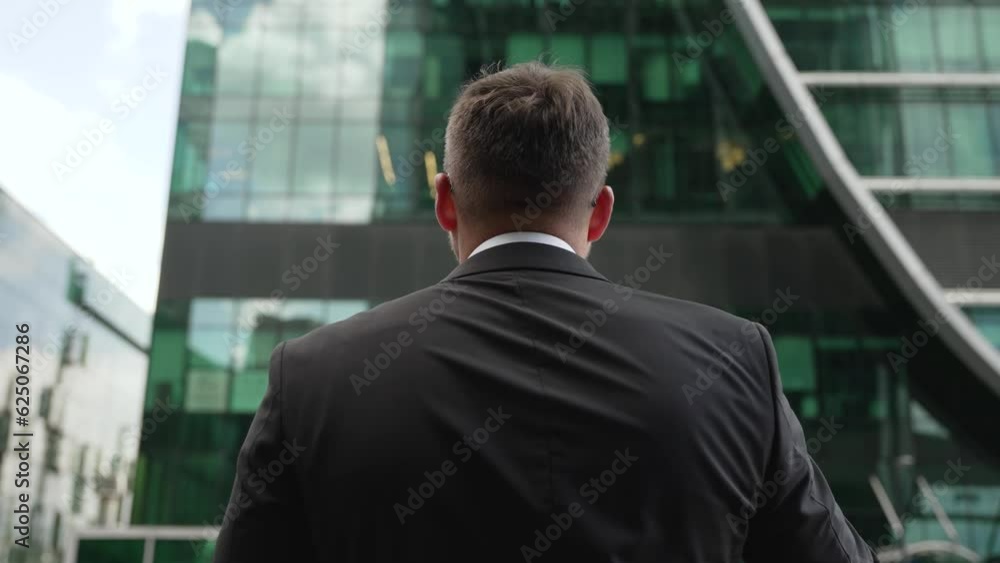 The businessman stands with his back and looks at the skyscraper. Rear view of a thoughtful businessman in a suit standing on the street with his hands in his pockets. View of the business district of The businessman stands with his back and looks at the skyscraper. Rear view of a thoughtful businessman in a suit standing on the street with his hands in his pockets. View of the business district of