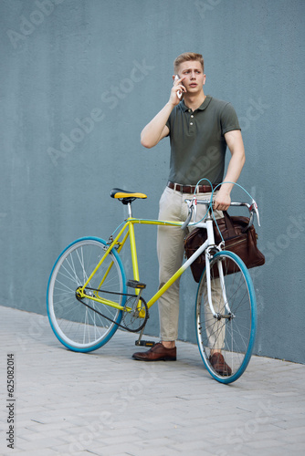 Handsome, concentrated, stylish businessman in smart casual clothes standing outdoors with bike and talking on mobile phone. Concept of business, active lifestyle, fashion, youth, ecology