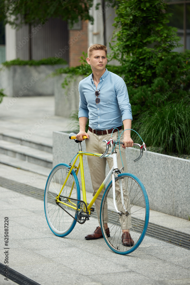 Young handsome businessman, office worker in stylish smart casual clothes standing outdoors with bike. Beautiful urban background. Concept of business, active lifestyle, fashion, youth, ecology