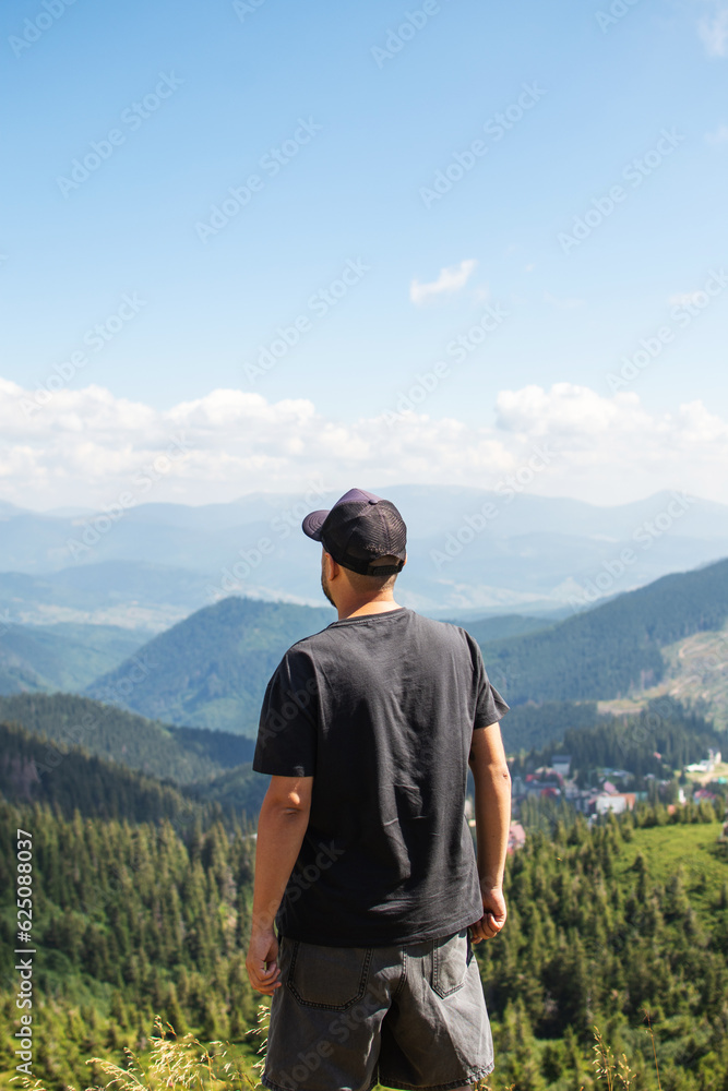 Back view of young male traveler wearing cap standing alone in summer woods and looking at the mountain hills. Tourist guy enjoying the coniferous forest scenery and mountain landscape