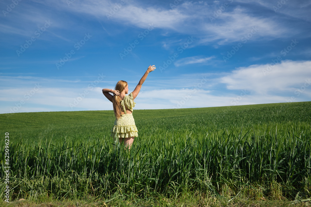 Young, beautiful, blonde woman in a yellow dress, with one arm raised in the middle of a green wheat field, seen from the back, alone, calm and peaceful. Concept tranquility, calm, fields, meadows. Young, beautiful, blonde woman in a yellow dress, with one arm raised in the middle of a green wheat field, seen from the back, alone, calm and peaceful. Concept tranquility, calm, fields, meadows.