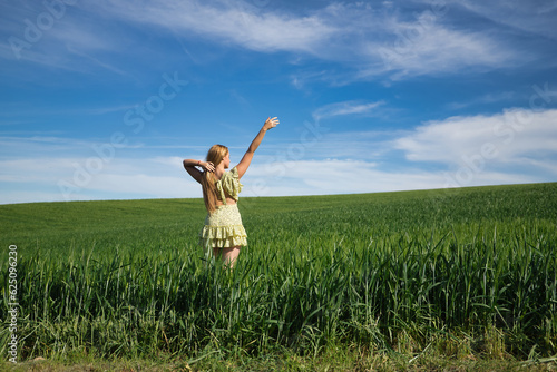 Young, beautiful, blonde woman in a yellow dress, with one arm raised in the middle of a green wheat field, seen from the back, alone, calm and peaceful. Concept tranquility, calm, fields, meadows.