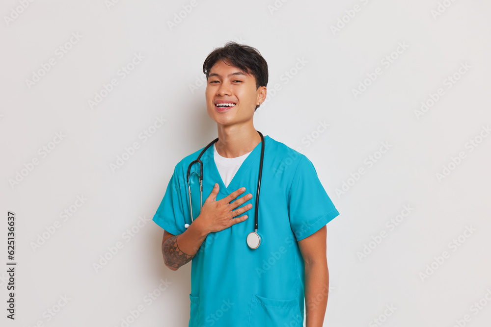 Young smiling male doctor in medical uniform poses on white background, looks into camera with hand on his chest, professional people concept, copy space Young smiling male doctor in medical uniform poses on white background, looks into camera with hand on his chest, professional people concept, copy space
