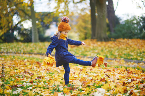 Adorable preschooler girl walking and kicking fallen leaves in autumn park in Paris, France