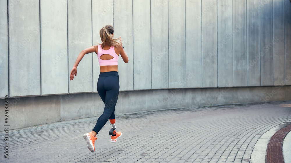 On the way to success.Back view of young disabled woman with leg prosthesis in comfortable sports clothing is running outdoors along the street On the way to success.Back view of young disabled woman with leg prosthesis in comfortable sports clothing is running outdoors along the street