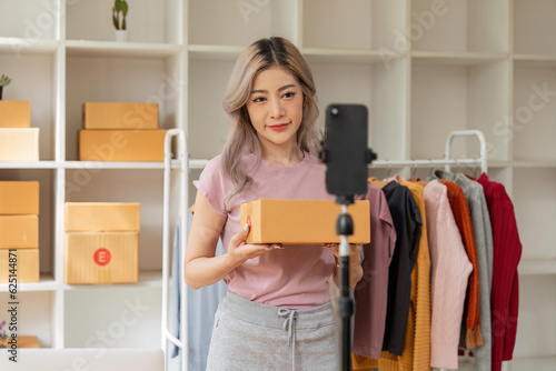 Asian beautiful woman sell clothes online live streaming at home. Young female use mobile phone video call shows goods to customer and present detail. Remote buying and purchase shopping concept