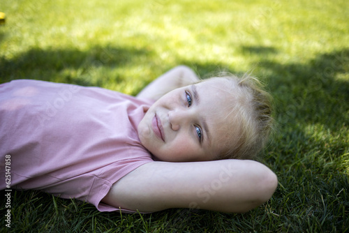 Portrait of a cute girl in the park, top view. Happy child playing outdoors in summer. The child is lying on the green grass. Earth Day and the concept of ecology. © evgeniia_1010