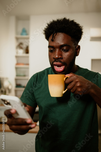 A shocked african american man is holding a mug while reading a disturbing message on the phone at home.