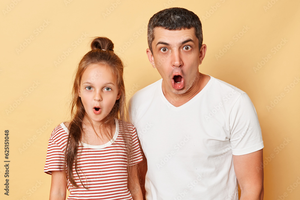 Shocked scared father and daughter wearing casual t-shirts standing isolated over beige background looking at camera with opend mouths and big eyes shock content. Shocked scared father and daughter wearing casual t-shirts standing isolated over beige background looking at camera with opend mouths and big eyes shock content.
