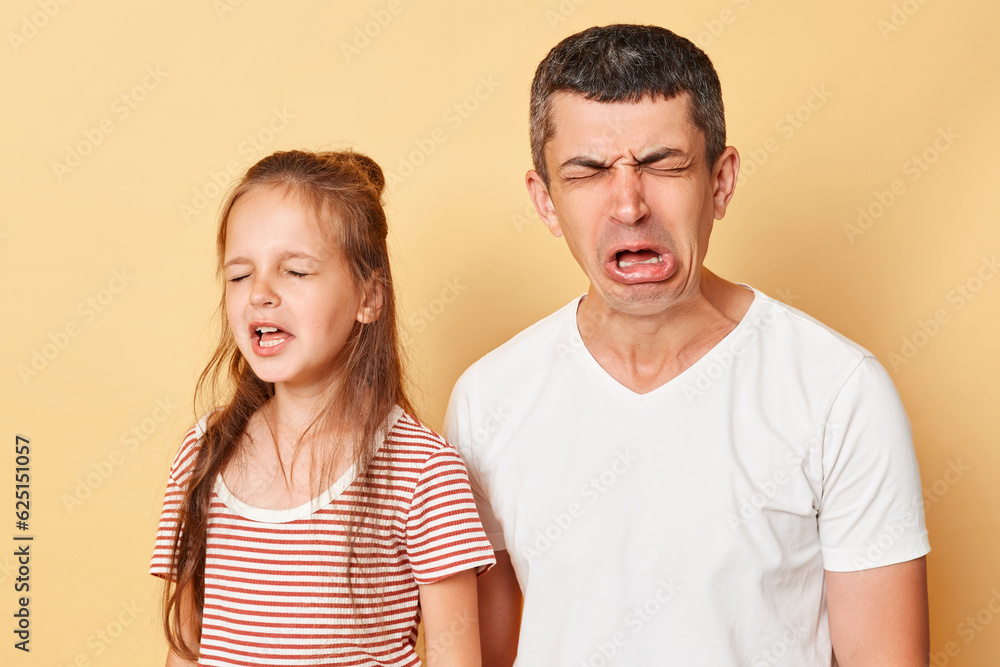 Funny father and daughter wearing casual t-shirts standing isolated over beige background crying grimacing expressing sadness and sorrow hearing bad news. Funny father and daughter wearing casual t-shirts standing isolated over beige background crying grimacing expressing sadness and sorrow hearing bad news.
