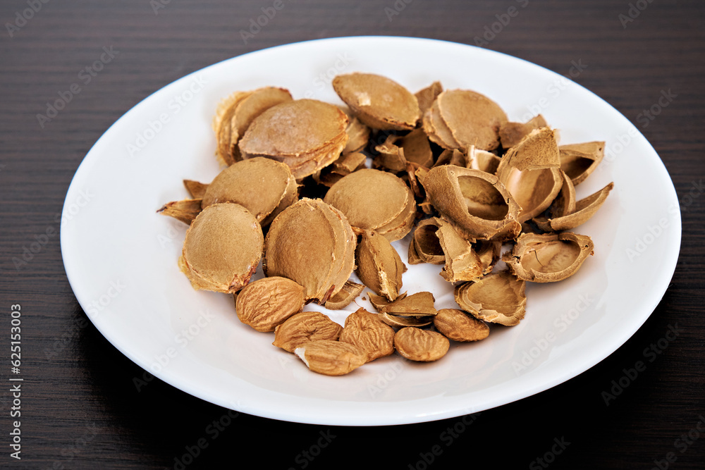 Apricot kernels and stones lie on a white plate on a dark background. View from above. There is space for text. Close-up Apricot kernels and stones lie on a white plate on a dark background. View from above. There is space for text. Close-up