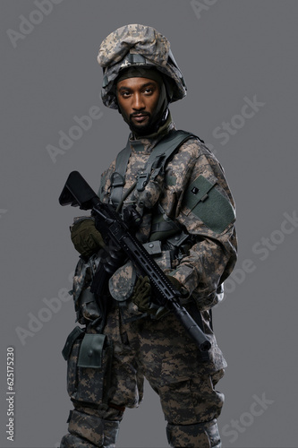 Dark skinned soldier in NATO uniform and helmet poses with a serious expression on a plain grey background