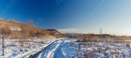 Winter landscape. Road covered with snow and a hill with tall dry grass on a sunny day.