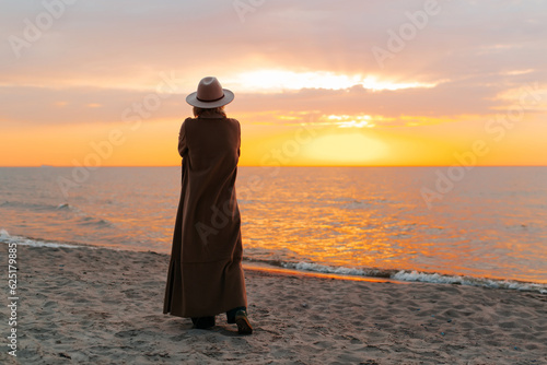 Lonely pensive woman walking at sunset by sea, enjoying nature. Rear view of unrecognizable stylish senior lady standing alone on seashore against yellow sky, copy space