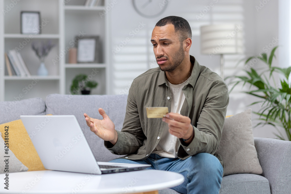 Financial problems. Upset African American young man sitting at home in front of laptop, looking worriedly at screen, holding credit card and spreading hands Financial problems. Upset African American young man sitting at home in front of laptop, looking worriedly at screen, holding credit card and spreading hands