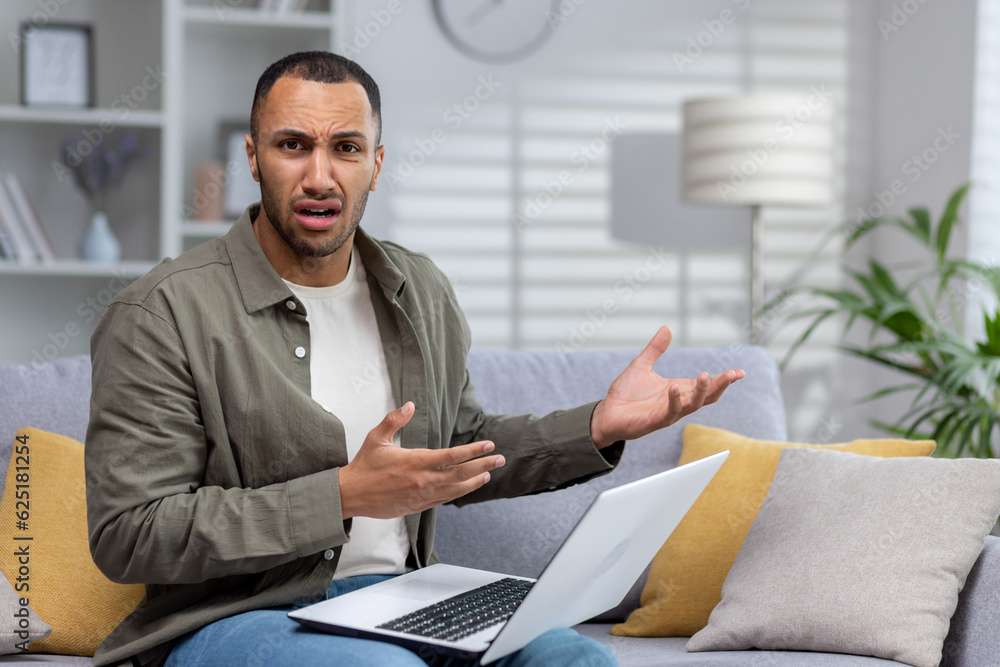 African American young man businessman, freelancer working from home on laptop. He looks worriedly and angrily at the camera, gesturing inexplicably with his hands African American young man businessman, freelancer working from home on laptop. He looks worriedly and angrily at the camera, gesturing inexplicably with his hands